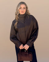 Woman wearing a dark brown poncho holding a brown leather handbag against a beige background