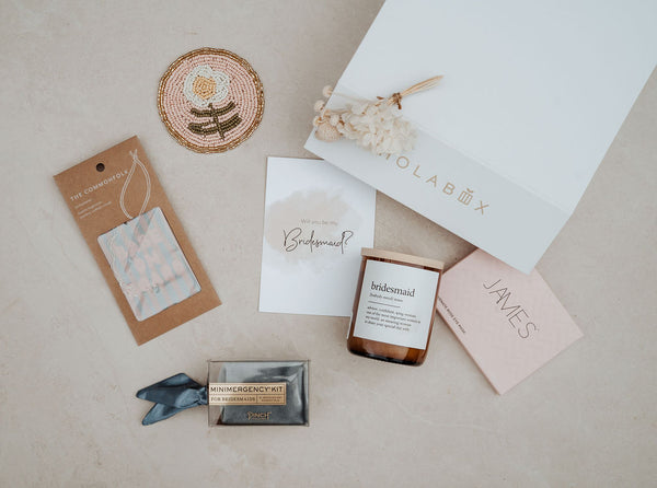 Collection of bridesmaid gifts including a box, card, and small items on a neutral background