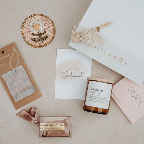 Collection of bridesmaid gift items including a box, card, and small bottles on a beige surface.