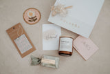 Collection of bridesmaid gifts including a candle, card, and box on a neutral background.