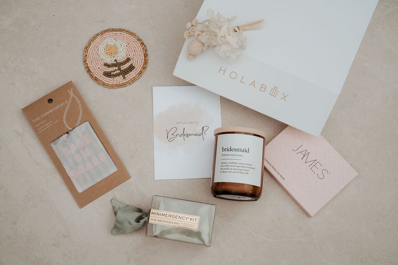 Collection of bridesmaid gifts including a candle, card, and box on a neutral background.