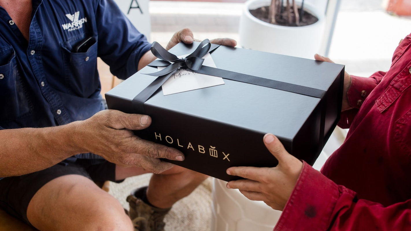 A black HOLABOX gift box tied with a black ribbon and bow, being handed over from a man in a blue shirt to a woman in a red jacket. The brand name 'HOLABOX' is printed in gold on the side of the box.