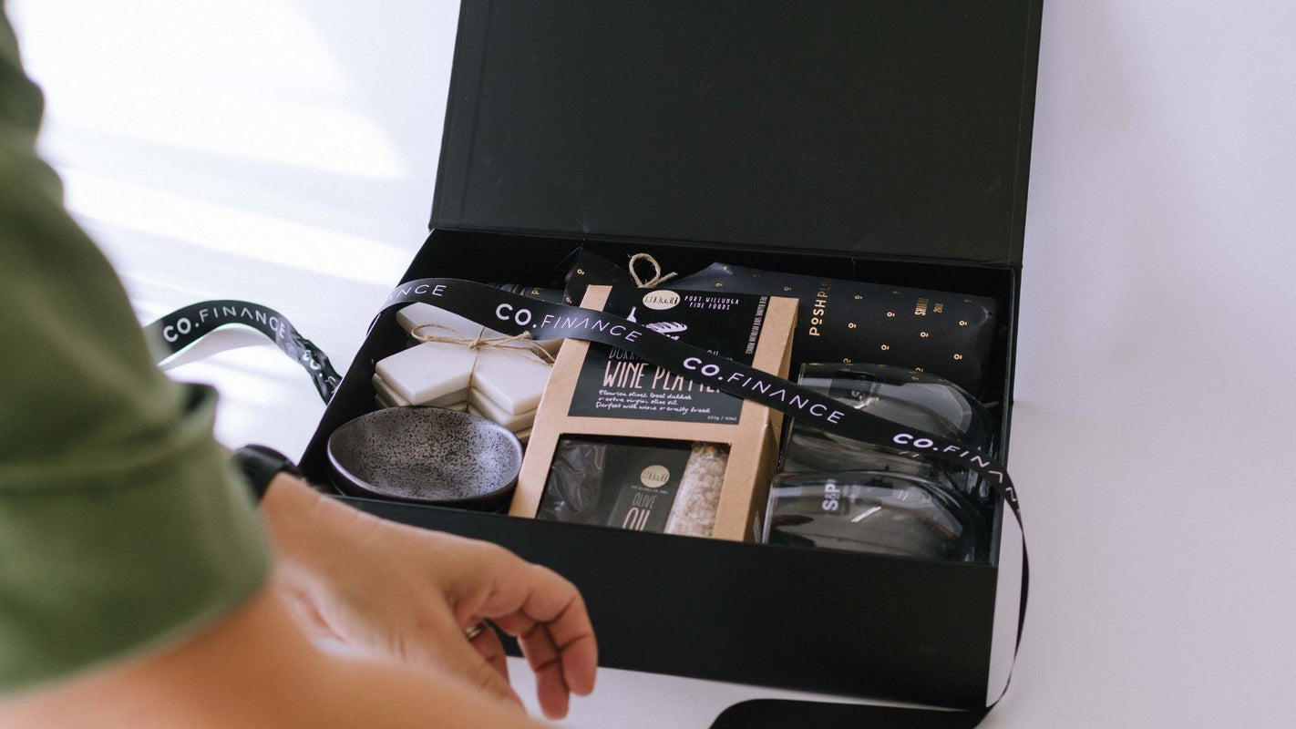 An open black gift box containing a curated collection of products from Co. Finance, including wallets, notebooks, and accessories, with a person's hand and arm visible on the left.