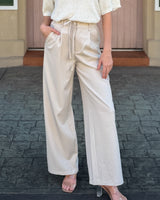 A person wearing high-waisted, wide-leg satin-like pants in a light cream or beige color, paired with a textured cream top and clear high-heeled sandals, standing outdoors on a dark paved surface.
