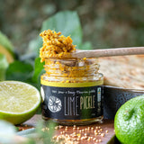 Jar of lime pickle with a wooden spoon, surrounded by limes and seeds on a wooden board.