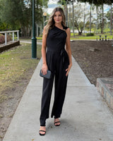 A woman stands on a concrete pathway in a park wearing a sleeveless black jumpsuit with a draped neckline, a tied waist belt, and wide legs. She is holding a small black clutch and wearing black heeled sandals.