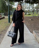 A woman stands on a paved pathway in a park, wearing a black draped long-sleeve top and matching wide-leg pants, holding a black structured handbag.