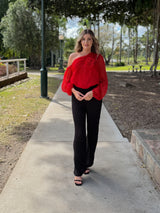 Woman wearing a red one-shoulder viscose top with a floral appliqué and voluminous sleeve, paired with black trousers.