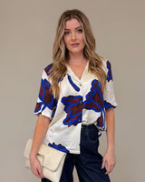 Woman wearing a patterned blouse and dark pants against a beige background
