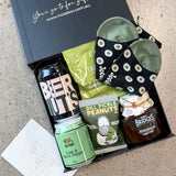 A curated gift box from HOLA BOX, opened to reveal a curated assortment of gourmet snacks and drinks on a speckled stone surface. The box contains a black can of BIER UT, a jar of lime and lemon sparkling water, a bag of dill pickle peanuts, and a small jar of Bridges Royal Jelly.