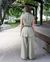 Back view of a sage green belted wrap vest with notched lapels, paired with matching wide-leg trousers in a park setting.