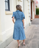 Back view of an indigo denim midi dress with a belted waist, short sleeves, and pockets, worn on a city sidewalk.