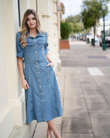 Woman wearing an indigo denim midi dress with a belted waist, button-down front, and pockets, standing on a sidewalk.