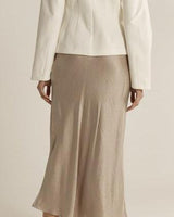 Taupe midi-length bias cut skirt in smooth satin, paired with a white blazer for an elegant silhouette.