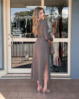 Woman wearing a brown polka dot midi dress with long sleeves, ruched detailing, and a side slit, paired with clear heels.