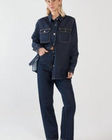 Aveline dark denim shirt with silver buttons, chest pockets, and a curved hem worn with matching jeans.