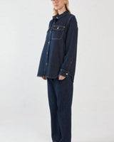 Dark blue denim shirt with chest pockets, silver buttons, and a curved hem worn with matching denim trousers.