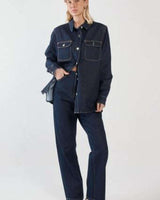 Aveline dark denim shirt with silver buttons, chest pockets, and a curved hem worn with matching jeans.