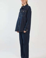 Dark blue denim shirt with chest pockets, silver buttons, and a curved hem worn with matching denim trousers.