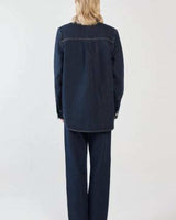 Back view of a woman wearing a dark blue denim shirt with a curved hem and matching denim trousers.