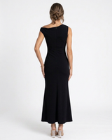 A woman viewed from the back wearing a sleeveless black off-the-shoulder maxi dress with structured shoulders and a fitted silhouette, paired with clear high-heeled sandals against a plain light gray studio background.