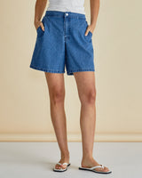 Relaxed-fit blue denim shorts with a flat front, zip fly, and side pockets worn with a white tank top.