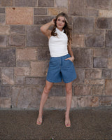 Woman wearing relaxed-fit blue denim shorts with a flat front, pockets, and belt loops against a stone wall.
