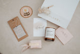 Collection of bridesmaid gifts including a candle, card, and other items on a neutral background.
