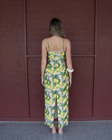 Back view of a lemon-print cotton blend maxi dress with spaghetti straps and a back slit against a brown wall.