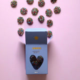 A light blue box of Sweeta Chocolate Covered Pretzels with a heart-shaped window, shown open with several pretzel rods covered in colorful sprinkles arranged above it on a solid pink background.