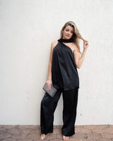 Woman wearing a black satin one-shoulder top with an integrated necktie and draped overlay paired with matching trousers.