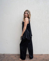 Woman wearing a black satin one-shoulder top with a draped overlay and integrated necktie, paired with matching pants.