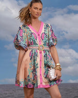 Dali mini dress in Cali pink with a V-neck, button-up front, puffed sleeves, and a patterned belt against a sky background.
