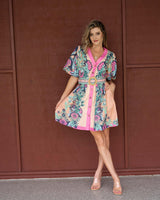 Woman wearing a pink and blue floral print linen mini dress with a V-neck, button-up front, and a woven belt.