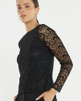 Model wearing a black Effie mesh top with a boat neckline, flocked floral pattern, and sheer long sleeves.