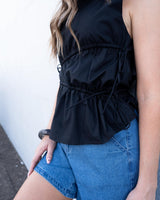 Black cotton sleeveless top with adjustable side ties and asymmetric gathering, styled with blue denim shorts.