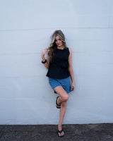 Black cotton sleeveless top with adjustable side ties and asymmetric gathering, paired with blue denim shorts.