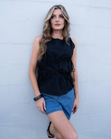 Black cotton sleeveless top featuring asymmetric gathering and adjustable tie sides, styled with denim shorts.