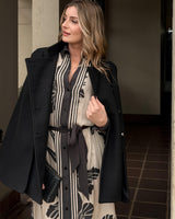Woman wearing a black wool-blend military cape with double-breasted buttons over a striped patterned dress.