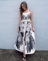 Woman wearing a cream and black linen-blend sundress with adjustable straps, pockets, and intricate floral print.
