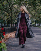 A woman wearing a long grey suede trench coat over a burgundy ruched maxi dress while walking outdoors.