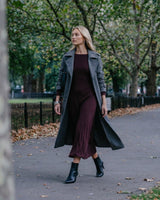 A woman wearing a charcoal grey suede trench coat over a long burgundy dress while walking in a park.