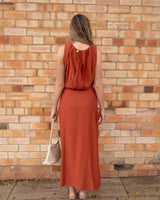 Back view of a rust-coloured sleeveless midi dress with a button-back detail, worn against a brick wall.