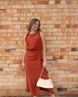 Woman wearing a sleeveless rust-coloured midi dress with a gathered waist and side slit against a brick wall.