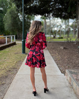 Woman wearing a long-sleeve, red and black floral print mini dress with a tie waist and ruffle hem outdoors.