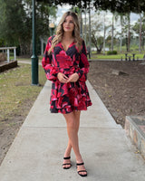 Woman wearing a black and red floral print mini dress with long sleeves, a wrap-style V-neck, and a ruffled hem.