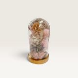 House of Holabox floral cloche with preserved pink and neutral dried flowers inside a glass bell jar on a gold base.
