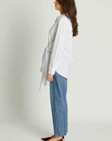 Woman wearing a white 100% cotton wrap-style shirt with an adjustable side tie, paired with blue denim jeans.
