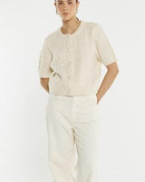 Cream cable knit short-sleeve cardigan with self-covered buttons and ribbed trim worn with matching trousers.