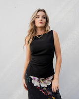 Sleeveless black Hannah apron top with boat neckline, side ruching, and peplum hem worn with a floral skirt.